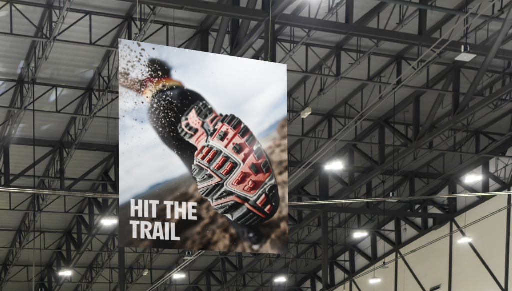 A hanging banner advertisement in a large convention space or warehouse. The ad shows trail running shoes. 